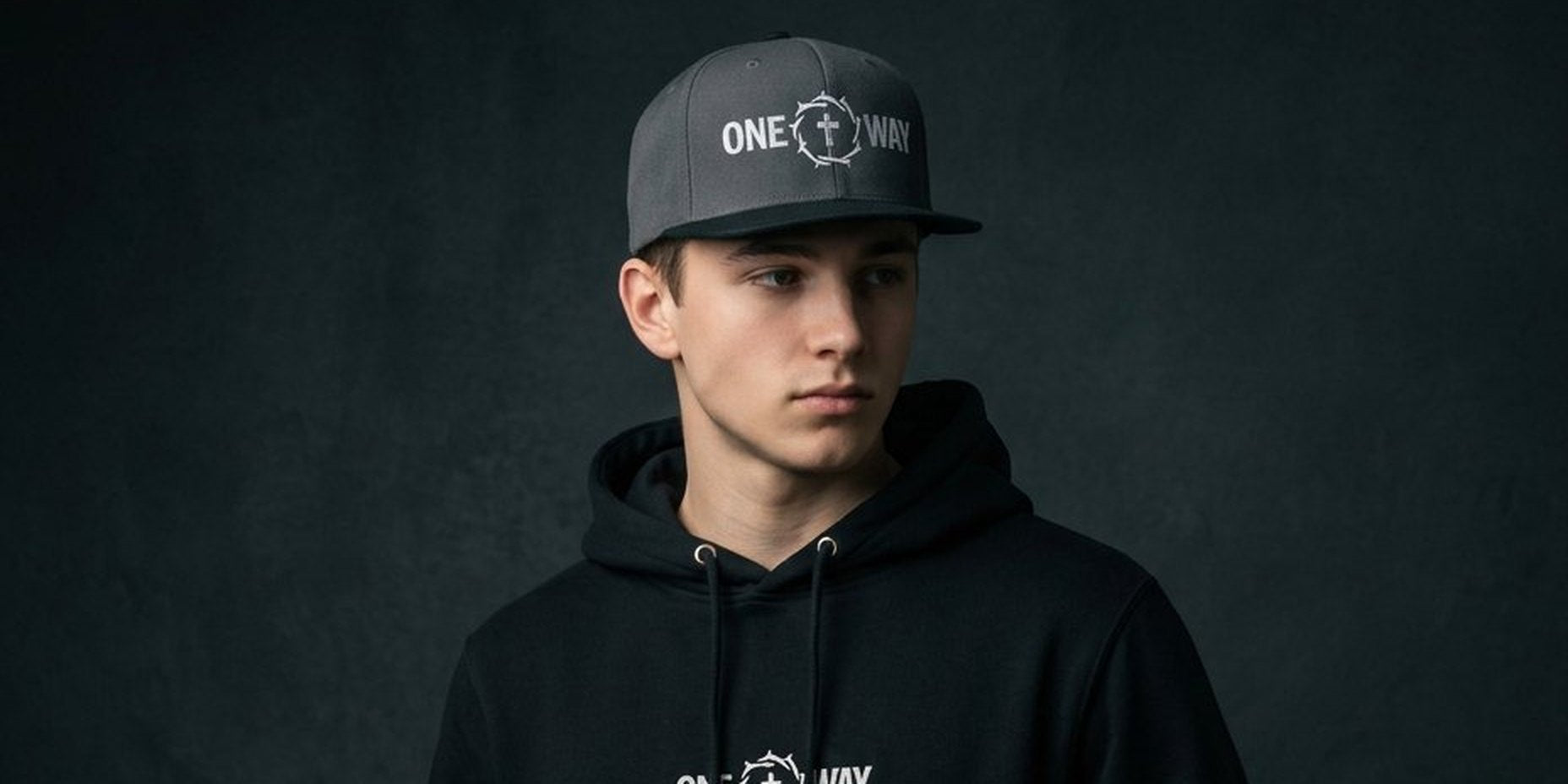 Image of Young man in a black One Way hoodie and cap against a dark studio wall