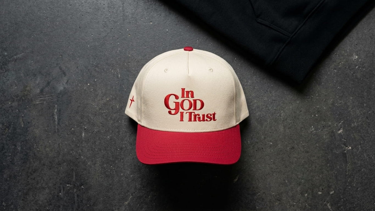 Cream In God I Trust cap with a red visor and red embroidery, on dark concrete beside a black hoodie