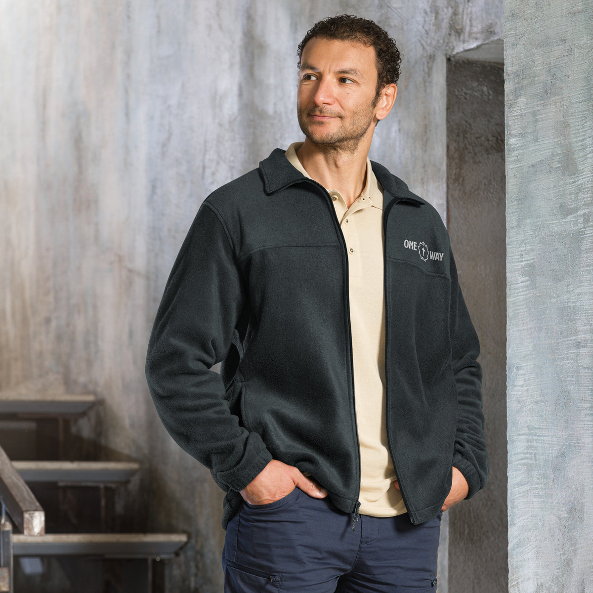 Man standing by a concrete wall wearing the One Way Columbia fleece jacket in black with the ONE WAY embroidery on the chest